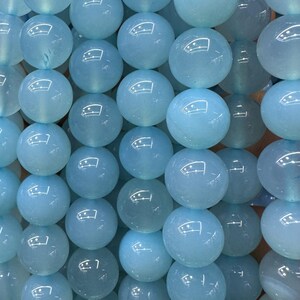 May include: A collection of light blue, round gemstone beads. The beads are smooth and polished, with a translucent quality. The beads are arranged closely together, creating a textured surface. The color is a soft, pale blue, reminiscent of aquamarine.