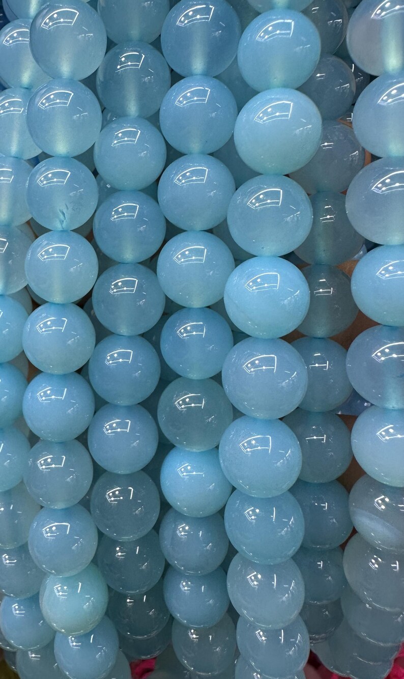 May include: A collection of light blue, round gemstone beads. The beads are smooth and polished, with a translucent quality. The beads are arranged closely together, creating a textured surface. The color is a soft, pale blue, reminiscent of aquamarine.