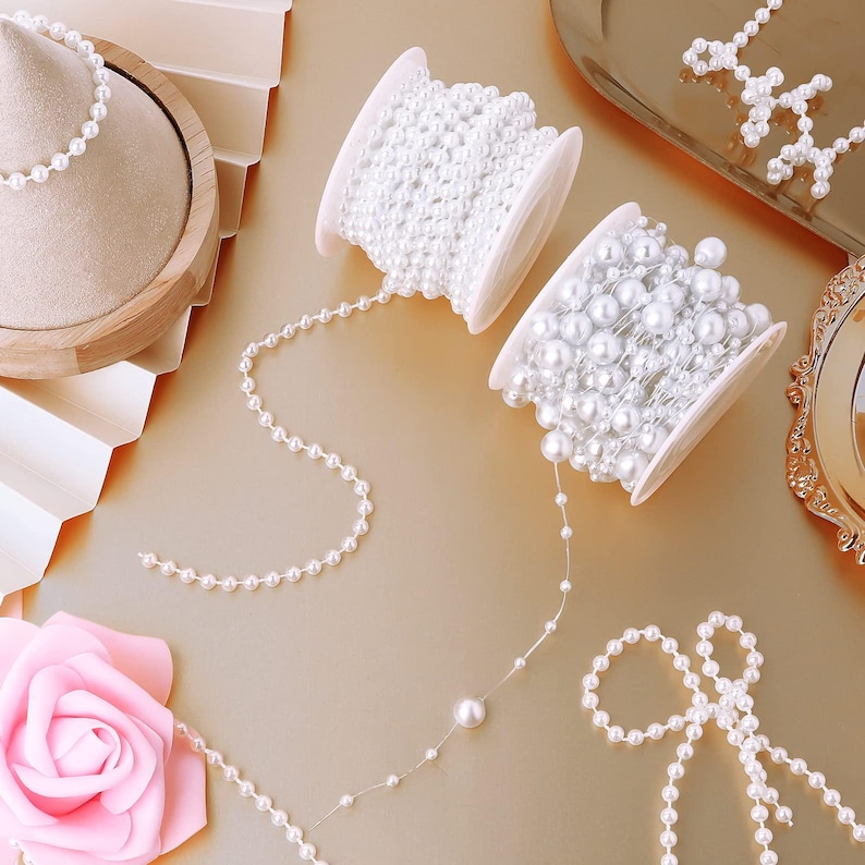 May include: White pearl bead strands and spools are displayed on a gold surface. The image features two spools of white pearl beads, a strand of pearls, and a pink rose. The pearls vary in size and are used for jewelry making or crafting.