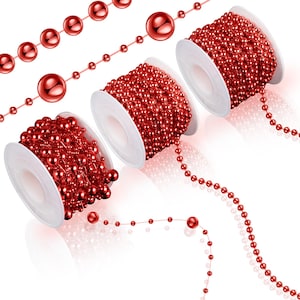 May include: Red beaded garland on white spools and loose strands. The garland features small, round beads and larger, spherical beads. The spools are arranged at different angles, showcasing the garland's texture and color. The beads have a glossy finish.