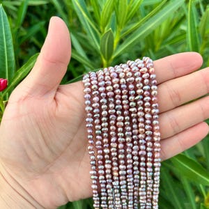 May include: A hand holds multiple strands of small, oval pearls. The pearls display a mix of pink, lavender, and silver hues. The background is a blur of green foliage and a few red flowers, creating a natural setting for the jewellery.