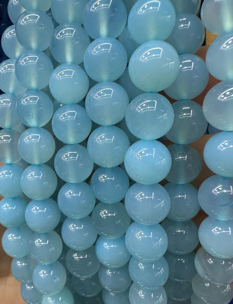 May include: Close-up of strands of light blue, round beads. The beads are smooth and have a glossy finish, reflecting light. The beads are strung together, creating a textured surface. The overall theme is jewelry making or crafting.