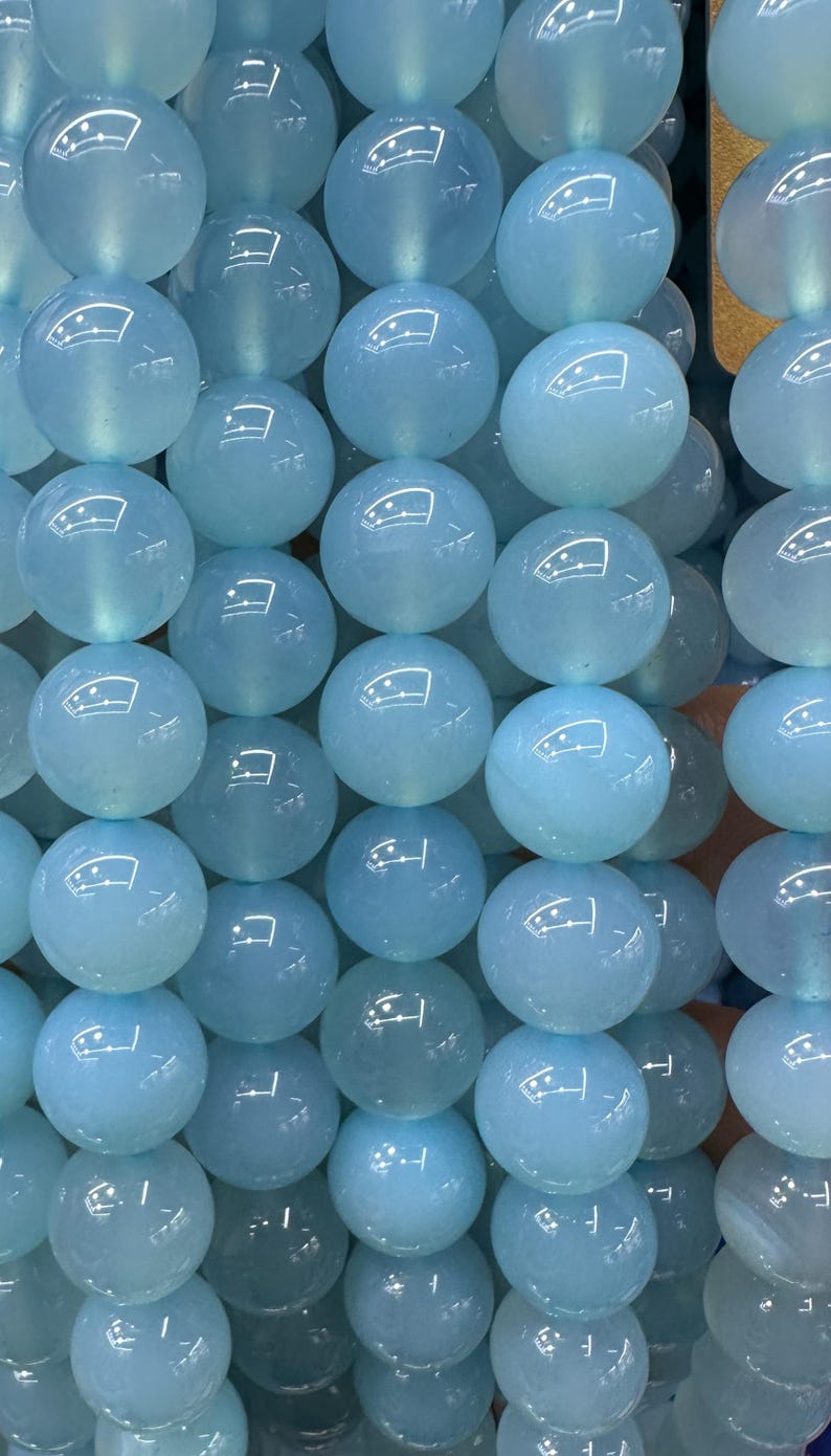 May include: Close-up of a collection of light blue, round beads. The beads have a glossy, reflective surface and appear to be strung together. The beads are a consistent size and shape, creating a uniform pattern. The overall theme is jewelry making or crafting.