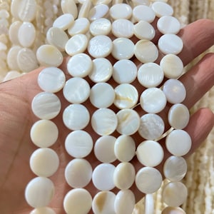 May include: Close-up of strands of white, round mother-of-pearl beads. The beads are flat and disc-shaped, with a pearlescent sheen. The beads are being held in a hand, with other strands visible in the background. These beads are ideal for jewelry making.