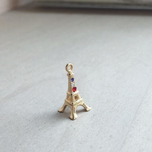 May include: A gold-colored Eiffel Tower charm with red, white, and blue gemstones. The charm is a popular souvenir and can be used for jewelry making.
