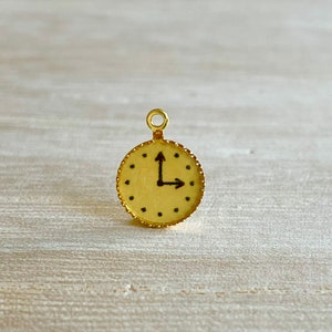 May include: A gold-coloured clock charm with a white face and black hands. The clock face has black markings for the hours and minutes.