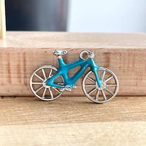 May include: A teal blue bicycle charm with silver wheels and a silver frame. The charm is small and would be perfect for a necklace or bracelet.