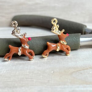 May include: Two gold-colored reindeer charms with red noses. The reindeer are brown and have gold antlers. The charms are on a wooden surface.