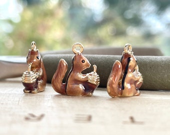 May include: Three gold-colored squirrel charms with brown enamel. Each squirrel is holding an acorn. The charms are on a wooden surface.