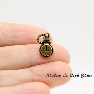 May include: A gold-toned metal charm shaped like an antique clock with a black face and Roman numeral markings. The charm has a small loop at the top for attaching to a necklace or bracelet.