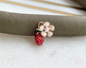 May include: A gold-tone charm featuring a red and green enamel strawberry with a white flower and a pearl center.