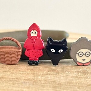 May include: Four small ceramic buttons featuring a red hooded girl, a black cat, a brown basket, and a woman with glasses. The buttons are all hand-painted and have a whimsical style.