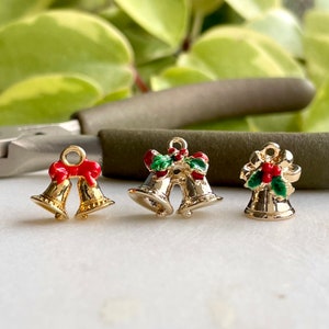 May include: Three gold-colored Christmas bell charms. One charm features a red bow, another has a green holly sprig with red berries, and the third charm has a green holly sprig with red berries and a gold bow.