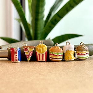 May include: Six colorful enamel charms in the shape of fast food items: a can of soda with "DRINK" written on it, a slice of pizza, a box of french fries, a hamburger, a beer mug, and another hamburger. The charms are all gold-toned and have a shiny finish.