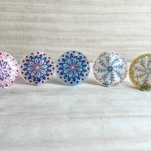 May include: Five round, decorative ornaments with intricate mandala designs. The ornaments are in shades of pink, blue, and yellow with gold accents.