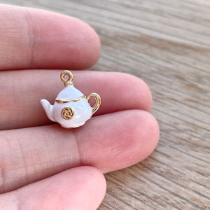 Pu&ograve; includere: Un ciondolo a forma di teiera bianca con dettagli dorati. La teiera ha un piccolo manico rotondo e un design dorato sul davanti.