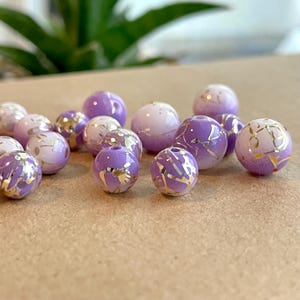 May include: A collection of purple and white glass beads with gold accents. The beads have a marbled effect and are perfect for jewelry making.