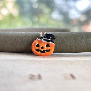 Könnte beinhalten: Ein kleiner, orangefarbener Kürbis-Anhänger mit einem schwarzen Hexenhut und silbernen Akzenten. Der Anhänger hat ein lächelndes Gesicht und ist wahrscheinlich für Halloween-Schmuck oder Bastelarbeiten gedacht.