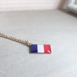 May include: A gold chain necklace with a pendant featuring the French flag. The pendant is a small, rectangular shape with the colors blue, white, and red.