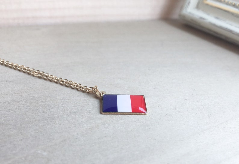 May include: A gold chain necklace with a pendant featuring the French flag. The pendant is a small, rectangular shape with the colors blue, white, and red.