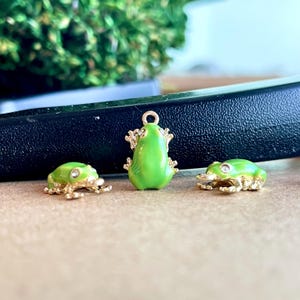 May include: Three green frog charms with gold accents. The frogs are in different poses, one is sitting, one is standing, and one is jumping.