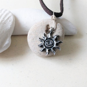 May include: A silver-colored sun pendant necklace with a detailed eye design at the center. The pendant is attached to a dark brown suede cord. The necklace is displayed on a light-colored stone.