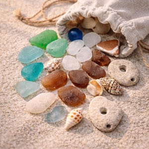 Könnte beinhalten: Eine Sammlung von Meeresglas in Grün-, Blau- und Brauntönen, zusammen mit Muscheln und kleinen Steinen, die aus einem beigen Kordelzugbeutel auf Sand fallen. Die Szene hat eine natürliche, strandähnliche Ästhetik.