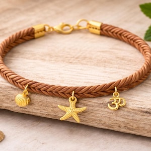 May include: Brown braided bracelet with gold charms. The charms are a seashell, starfish, and Om symbol. The bracelet has gold end caps and a clasp. The bracelet's circumference is approximately 18 cm.