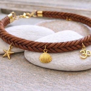 May include: A brown, braided leather bracelet with gold-coloured charms. The charms are a starfish, a seashell, and an Om symbol. The bracelet is displayed on white stones against a blurred background.
