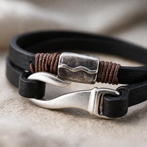 Puede incluir: Primer plano de una pulsera de cuero negro con cierre plateado. La pulsera presenta un diseño de doble vuelta, un adorno metálico decorativo y costuras marrones. El cierre es de tipo gancho y ojo.