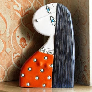 May include: A ceramic figurine of a person with a white face, bright blue eyes, and long dark hair. The figure wears an orange garment featuring a pattern of white polka dots and a white collar with small circular details along the neckline.