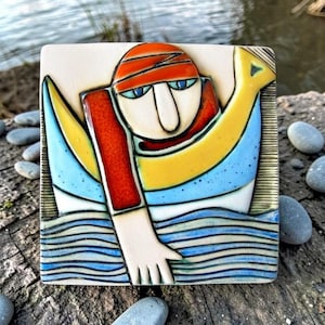 Pode incluir: Azulejo de cerâmica quadrado com um nadador num design estilizado. O nadador usa um boné laranja e mangas vermelhas, com olhos azuis e um nariz comprido. O fundo inclui elementos azuis e amarelos, com linhas de água azuis.