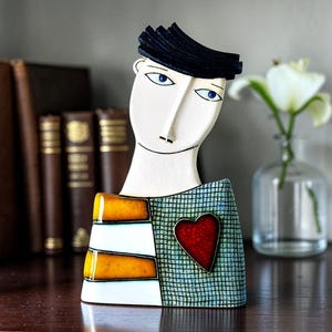 May include: A ceramic figurine of a person with a white face, blue eyes, and black hair. The torso features a yellow and white striped pattern on the left side and a light blue grid pattern on the right, accented by a prominent red heart.