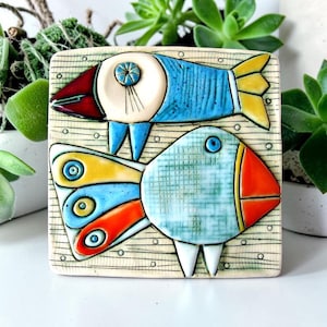 May include: A square ceramic tile featuring two stylized fish. The top fish is blue with a red head and yellow tail. The bottom fish is light blue with a textured grid pattern, an orange head, and a tail with blue, yellow, and orange segments.