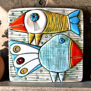 Può includere: Una placca da parete quadrata in ceramica con due pesci stilizzati. Il pesce superiore è giallo con coda blu e testa arancione. Il pesce inferiore è azzurro con un motivo a griglia, testa rossa e coda bianca con accenti circolari.