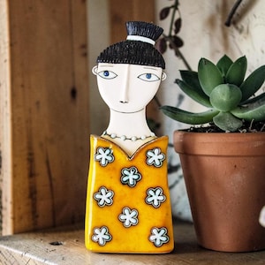 Könnte beinhalten: Keramikfigur einer Frau mit schwarzen Haaren und blauen Augen. Sie trägt ein gelbes Kleid mit weißen und blauen Blumenmustern. Die Figur steht auf einem Holzregal neben einer Topf-Sukkulente.