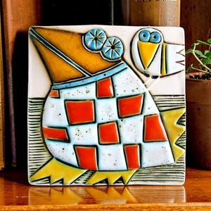 Könnte beinhalten: Eine quadratische Keramikfliese mit einer stilisierten Vogelillustration, die einen großen gelben Schnabel, blaue kreisförmige Augen und einen Körper mit weiß-orangem Karomuster aufweist. Der Vogel hat gelbe Füße und steht auf einer texturierten Oberfläche.