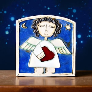 Op de afbeelding: Een keramieken engelenbeeldje met een blauwe achtergrond. De engel heeft gesloten ogen, krullend haar en witte vleugels. Het houdt een rood hart vast. Een halve maan en een ster staan op de achtergrond. Het beeldje staat op een houten oppervlak.