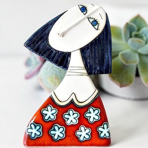 May include: A ceramic figurine of a stylised figure with a white face, blue eyes, and dark blue hair. The figure wears a red dress decorated with a repeating pattern of light blue and white flowers. The piece features black outlines throughout.