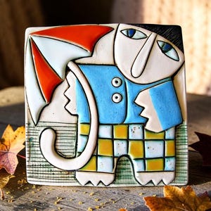 Può includere: Una piastrella quadrata in ceramica raffigurante una figura stilizzata che regge un ombrello rosso e bianco. La figura indossa una camicia azzurra con due bottoni bianchi e pantaloni a scacchi gialli e bianchi, delineati da contorni neri.