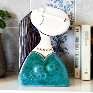 May include: A ceramic sculpture of a stylised figure with a white face, closed eyes, and long dark blue hair. The figure wears a beaded necklace and a textured, mottled teal green top. The piece has a smooth, glossy finish and a flat base.