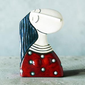 May include: A ceramic figurine of a stylised woman with closed eyes. The figure has a white head and neck, long blue hair, and a red dress with white polka dots. The figurine is on a grey surface.