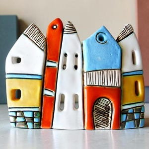 Könnte beinhalten: Eine Keramikskulptur, die eine Reihe von fünf stilisierten Häusern mit unterschiedlichen Dachformen darstellt. Die Strukturen sind in Weiß, leuchtendem Orange, Gelb und Hellblau bemalt und weisen strukturierte Details wie Fenster, Türen und Karomuster auf.