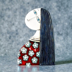 May include: A ceramic figurine of a stylized woman with long, dark blue hair. The figure has a white face with blue eyes and a red dress adorned with white and light blue flowers. The figurine has a unique, artistic design.