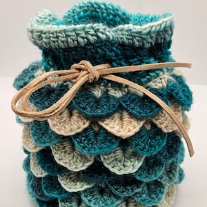 May include: A handmade crochet pouch in shades of teal, blue, and cream, resembling dragon scales. The pouch has a drawstring closure made of a light brown leather cord. The pouch is approximately 5 inches tall.