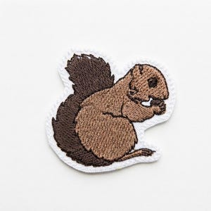 May include: An embroidered patch featuring a brown squirrel with a dark brown tail, holding a small nut in its paws. The squirrel is depicted in profile, rendered with detailed thread work in shades of light and dark brown, finished with a white border.