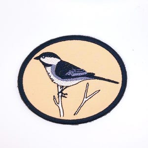 May include: A black and white embroidered bird patch on a beige background. The bird is perched on a branch and is surrounded by a black circle.