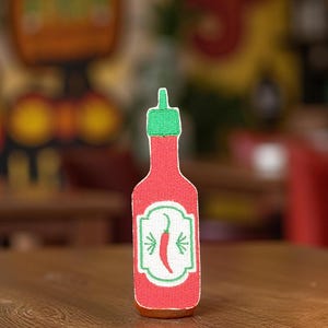 May include: A red embroidered patch in the shape of a hot sauce bottle. The bottle has a green cap and a white label with a red chili pepper and green leaves. The patch is on a wooden table.