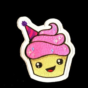 Peut inclure: Un patch de cupcake en feutre blanc avec du glaçage rose, un chapeau de fête violet et rose et un visage souriant. Le glaçage a des accents de paillettes.
