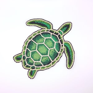 May include: An embroidered patch shaped like a sea turtle. The turtle features a shell with a geometric pattern in shades of bright green and dark green, outlined in a light beige thread. The flippers are dark green with bright green accents.